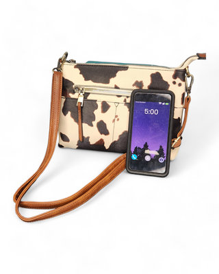 A cow print crossbody bag with a brown and cream cowhide pattern made from faux leather, featuring a rich brown strap and detailed stitching.