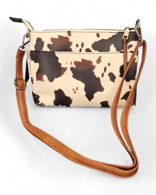 A cow print crossbody bag with a brown and cream cowhide pattern made from faux leather, featuring a rich brown strap and detailed stitching.