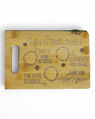 A bamboo Santa treat board engraved with placeholders for milk, cookies, and reindeer snacks, featuring whimsical text and snowflake designs.