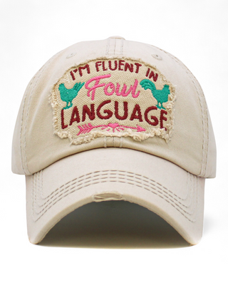 A distressed ivory baseball cap featuring an embroidered patch with the text "I'm Fluent in Fowl Language" and chicken graphics, providing a fun and stylish accessory.