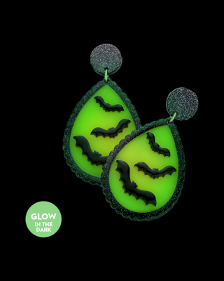 Glow-in-the-dark earrings featuring a teardrop design with vibrant green background and three black bat silhouettes in each earring. The earrings are outlined with a sparkling dark glitter and attached to glitter-covered discs,