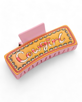 Rectangular hair clip with a 'Cowgirl' inscription set against a peach background, surrounded by a detailed floral pattern and housed in a pink clip frame