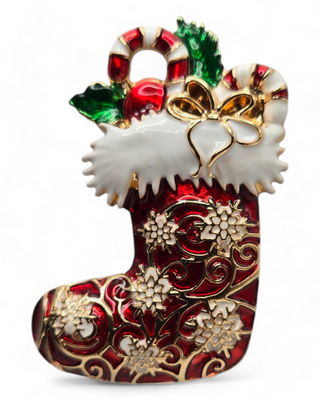 A Christmas stocking-shaped brooch with a red enamel finish, detailed with gold accents, white trim, and festive candy canes and holly leaves. The brooch is decorated with intricate patterns and holiday-themed designs.