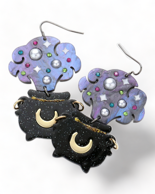 A pair of earrings featuring lavender clouds adorned with colorful gems and stars, attached to black cauldrons with crescent moons,