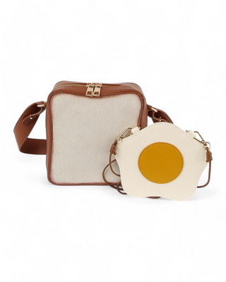 A toast-shaped crossbody bag in brown and cream paired with a small fried egg-shaped bag featuring a yellow yolk detail, both with gold-tone hardware and adjustable straps.