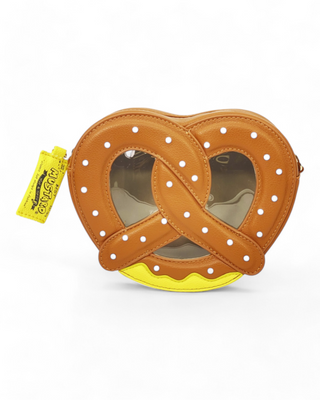 A caramel brown pretzel-shaped crossbody bag with white polka dot salt accents, a yellow mustard detail at the bottom, a transparent window center, and a detachable gold chain strap. One image shows the bag being worn with a white dress for a stylish look.